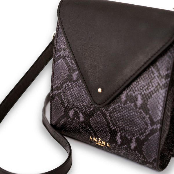 Amana Maggie Bag upclose angled front view. Black smooth leather bag with front panel black & grey embossed python print. Front flip opening with pin closure, gold zip closure at back, cellphone pocket and zip pocket in front interior, and cross body adjustable strap.
